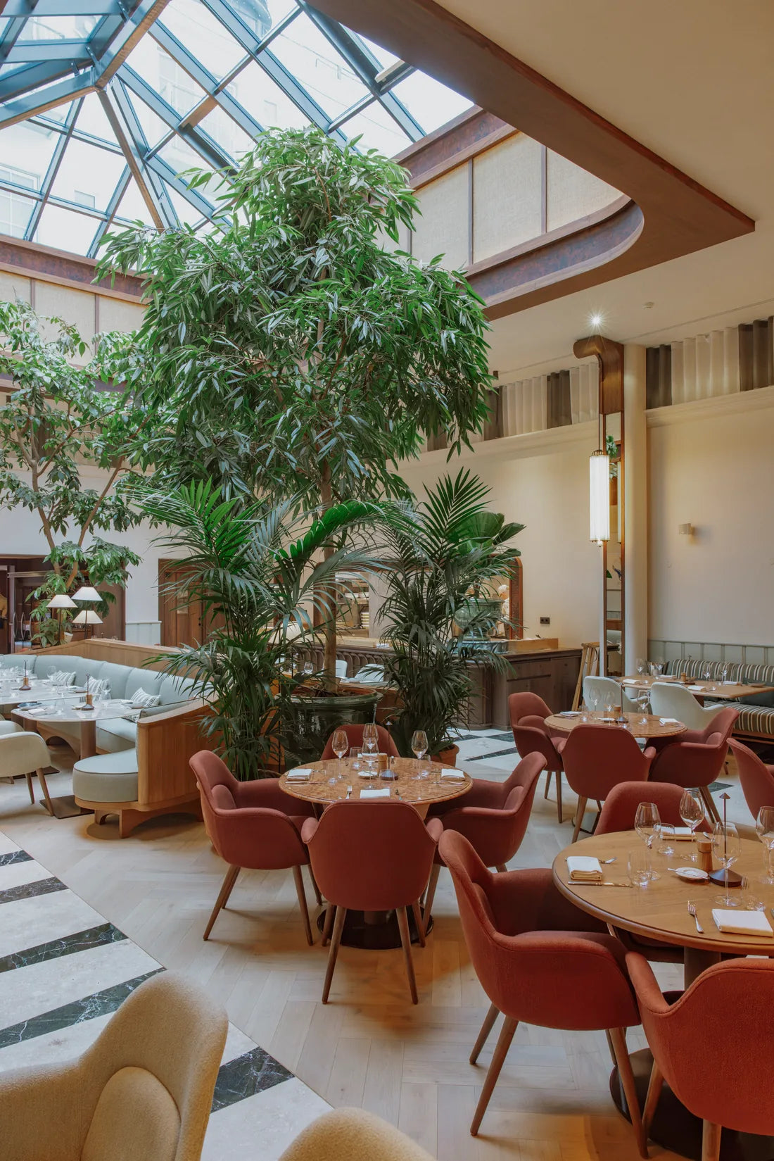 Dining tables, rust-coloured chairs and tall plants under the glazed roof at CORI Hornbæk.