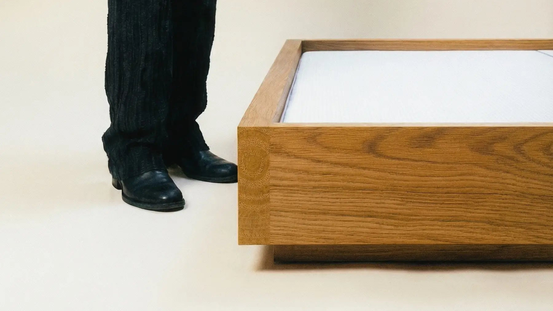 Person wearing a black outfit standing next to a wooden bed frame on a plain background