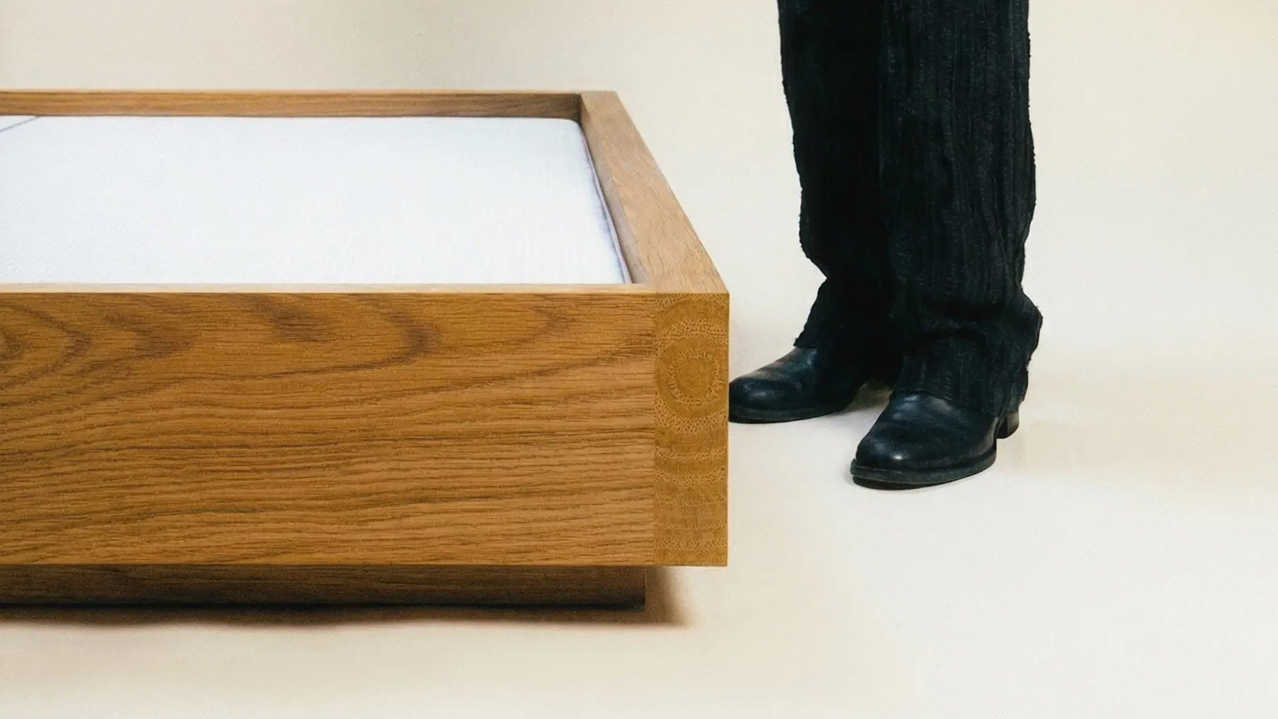 Person wearing a black outfit standing next to a wooden bed frame on a plain background